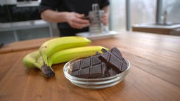 Schokolade in einem Schälchen auf dem Tisch, dahinter liegen vier Bananen. 