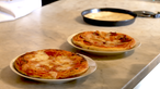 Zwei gebackene Pizzen auf Tellern, dahinter ungebackener Pizzateig auf einem Blech.