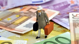 Das Bild zeigt eine miniatur Puppe mit Koffern, die auf Euroscheinen steht.