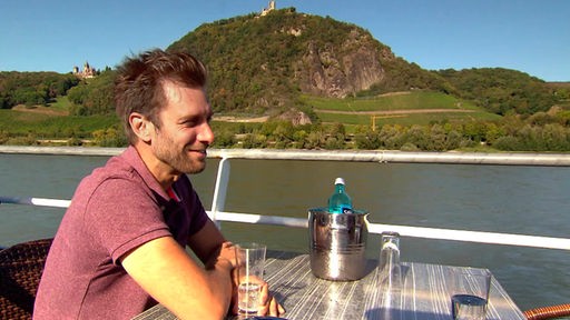 Daniel Aßmann sitzt an der Reling eines Rheinschiffs mit Blick auf den Drachenfels und die Weinberge. 