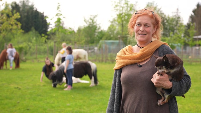 Eine rothaarige Frau hält einen kleinen Hund im Arm, im Hintergrund sind Menschen mit Ponys