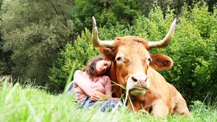 Eine junge Frau sitzt mit ihrem hellbraunen Rind kuschelnd auf einer Wiese 