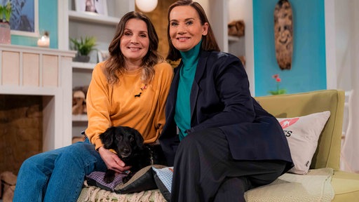 Simone Sombecki und Jenny Jürgens sitzen mit einem schwarzen Hund auf einem Sofa im Studio