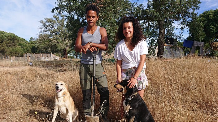 Nina und Stevo mit zwei Hunden auf einer sommerlichen Wiese in Portugal, im Hintergund Bäume und kleinere Gebäude. Stevo legt seine Hände über den Griff eines Spatens.