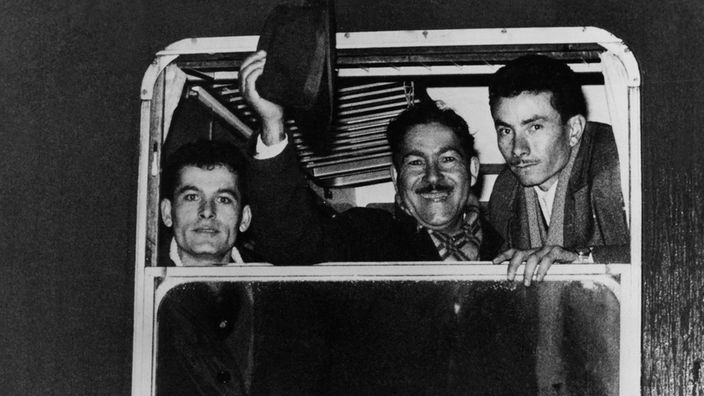 Ein Schwarzweiß-Foto: Drei Männer blicken aus einem Zugfenster, einer hebt seinen Hut