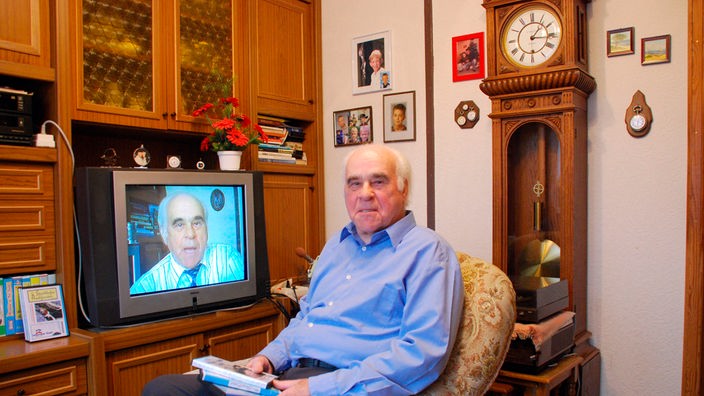 Ein älterer Mann sitzt in einem Sessel vor seinem Fernsehschrank und zeigt seine alten Aufnahmen aus der Vergangenheit.