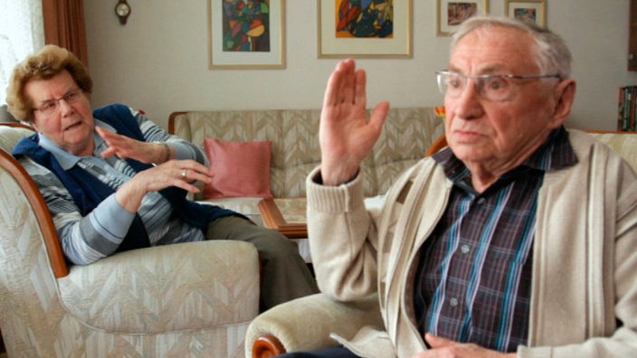 Ein älterer Mann und eine aältere Frau sitzen zuhause auf ihrer Sofa-Garnitur und sprechen über die Urlaubsreisen in der Vergangenheit.