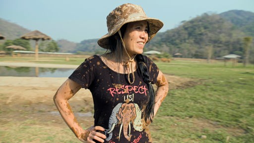 Die Elefantenretterin steht in einem Elefantenreservat, sie trägt einschlammbespritztes T-Shirt mit der Aufschrift Respect us und einem gezeichneten Elefantenkopf