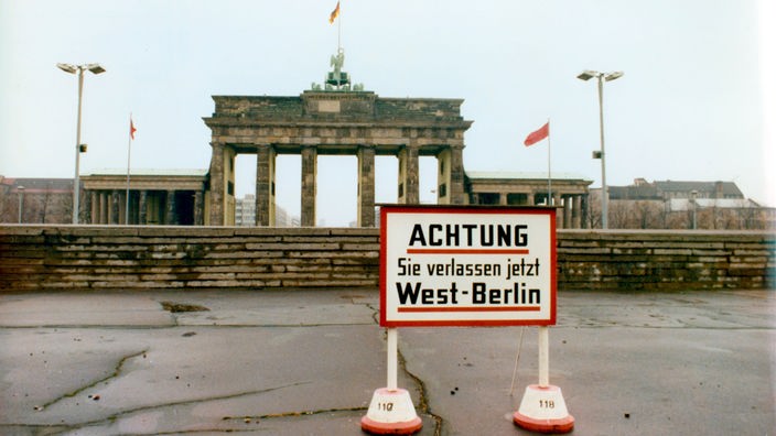 Blick auf die Berliner Mauer in den 1970er Jahren, im Hintergrund das Brandenburger Tor, im Vordergrund ein Schild mit der Aufschrift Achtung Sie verlassen jetzt West-Berlin