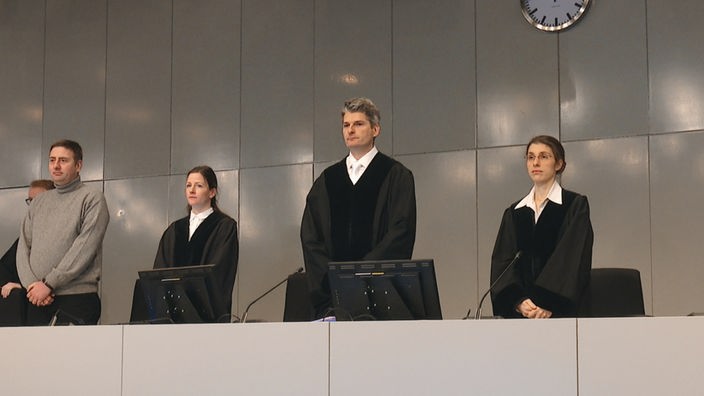 Ein Richter und zwei Richterinnen stehen im Gerichtssaal