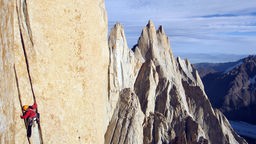 Blick auf eine Felswand, an der ein einzelner Bergsteiger emporklettert, im Hintergrund eine schneebedeckte Bergkette 