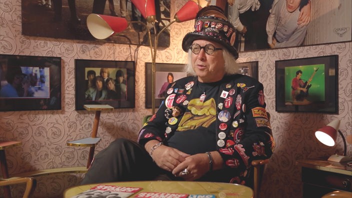 Ein Mann sitzt in einer Ecke umgeben von Bildern und Postern der Rolling Stones und trägt selbst diverse Merchandising-Artikel der Rockband.