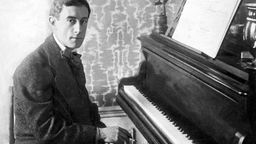 Der Komponist Maurice Ravel sitzt am Klavier und schaut in die Kamera. 