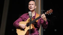 Ein Mann mit Glatze und weinrotem Hemd steht mit einer Gitarre vor einem Mikrofon und singt.