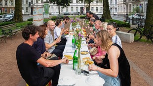Transurban Residency 23 in Krefeld, Straßenszene mit Menschen, die gemeinsam an einer großen Tafel essen