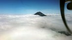 Blick aus einem Flugzeug auf einen Berggipfel, der sich aus den weißen Wolken in den blauen Himmel erhebt