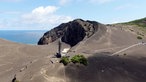 Ein einzelner Turm befindet sich inmitten einer graubraunen Vulkanlandschaft, im Hintergrund das Meer