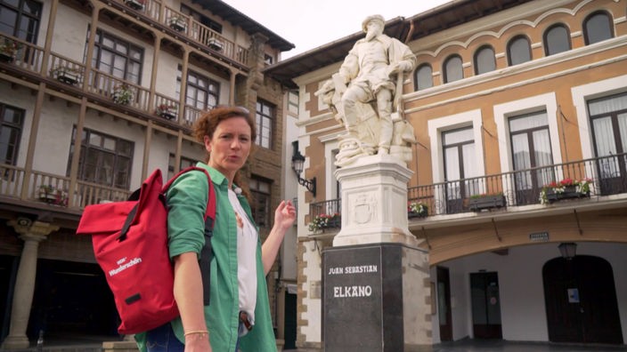 Anne Willmes steht vor der Statue des Seefahrers Elcano, im Hintergrund Stadthäuser