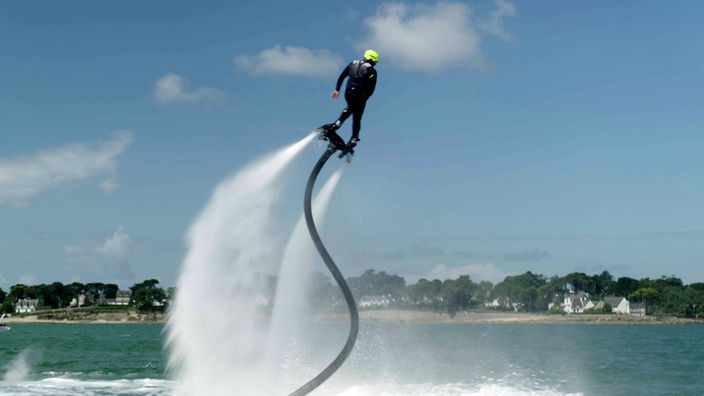 Ein Mann mit Helm steht auf einem Brett und wird von einem Schlauch mit Wasserkraft hoch geschleudert