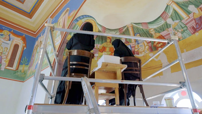 Zwei Nonnen stehen auf einem Gerüst und bemalen eine Kirchenkuppel