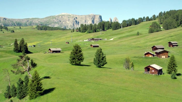 Blick über eine Bergwiese mit kleinen Hütten auf ein riesiges Felsmassiv