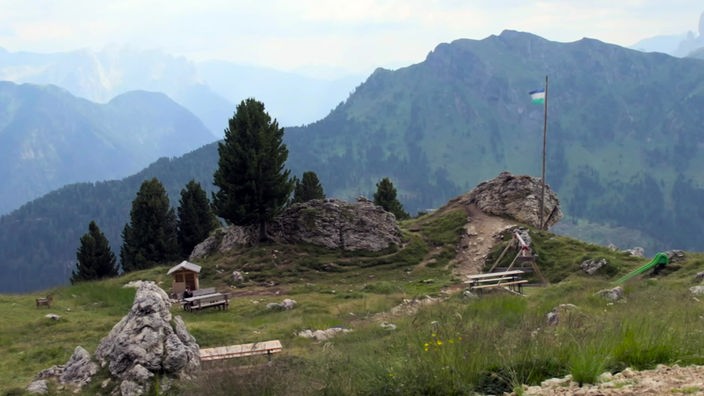 An einem überhängenden Felsen steht eine hölzerne Fahnenstange mit einer blau-weiß-grünen Fahne