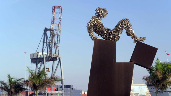 Stahlskulptur einer sitzenden Person auf Stahlblöcken, im Hintergrund Hafenkran