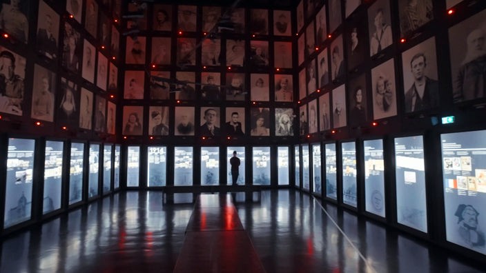 Einsamer Besucher steht in einem großen und sehr hohen Ausstellungsraum, dessen Wände bis in die Höhe mit historischen Portraitaufnahmen bedeckt sind