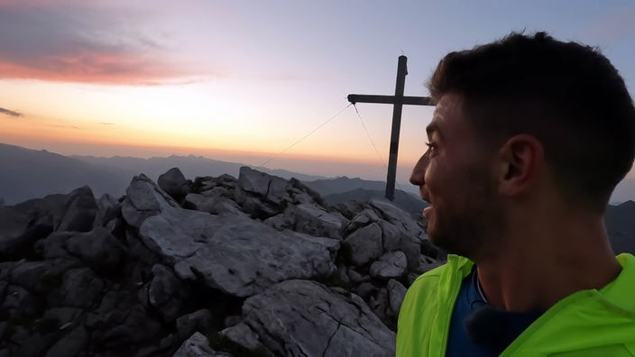 Ramon Babazadeh schaut lächelnd am Gipfelkreuz vorbei auf den Sonnenaufgang