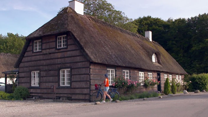 Tamina Kallert steht mit Fahrrad an einem Haus mit Reetdach und Reetwänden