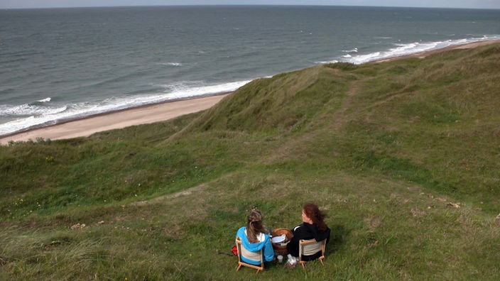 Tamina Kallert und eine Begleiterin sitzen auf Strandstühlen auf einer Düne und schauen aufs Meer