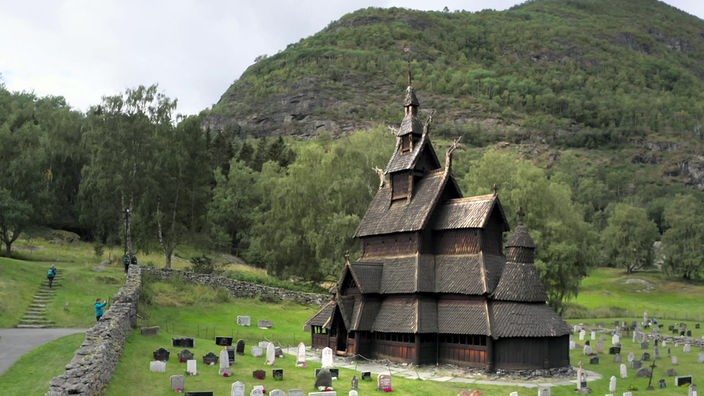 Holzkirche umgeben von Grabsteinen vor einem bewaldeten Hügel