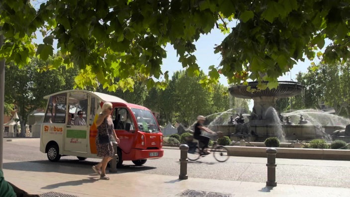 Ein Minibus fährt durch Aix-en-Provence. Im Hintergrund ein Springbrunnen