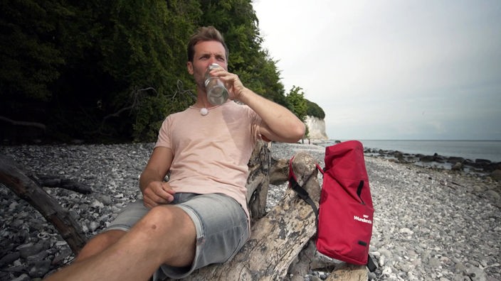 Daniel Aßmann sitzt am Kieselstrand und trinkt aus einer Flasche, im Hintergrund die Kreideküste