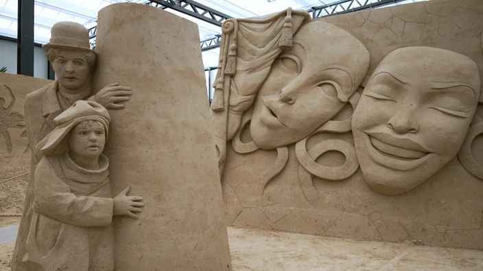Verschiedene Sandskulpturen wie Charlie Chaplin oder große Masken in einer Halle mit Glasdach