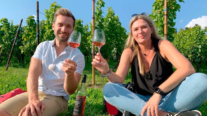 Daniel Aßmann und Sandra Polomski sitzen mit Weingläsern in der erhobenen Hand vor Weinstöcken