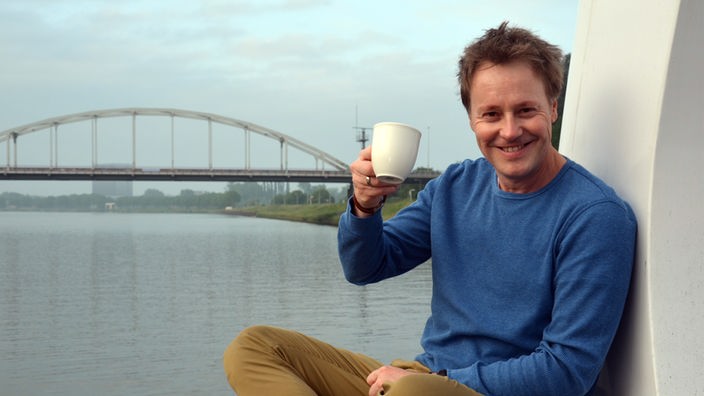 Stefan Pinnow winkt mit einem Kaffeebecher in die Kamera,im Hintergrund eine weiße Bogenbrücke