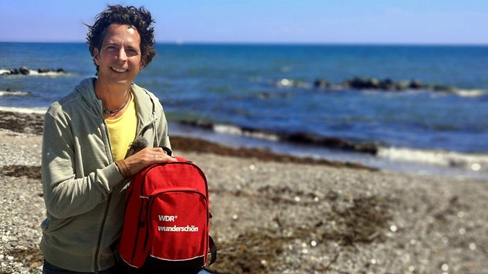 Moderator Jo Hiller steht mit dem roten Rucksack an der Ostseeküste