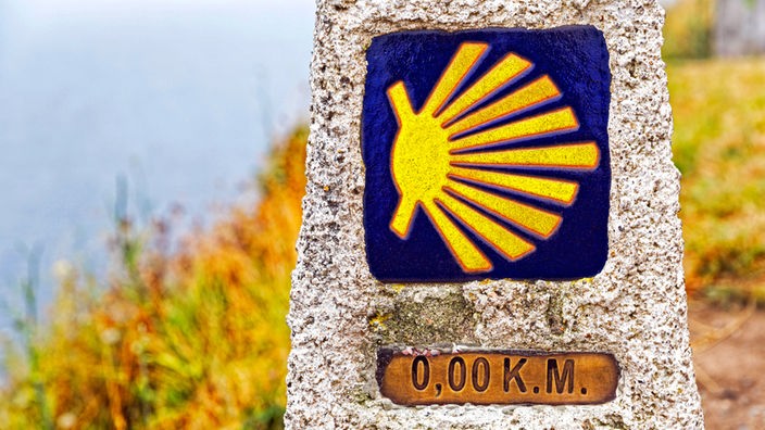Das Zeichen der Jakobsmuschel auf einem Stein und der Angabe "Kilometer Null"