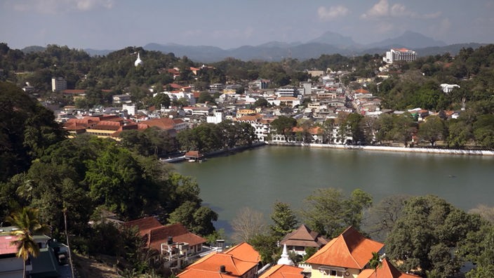 Die alte Königsstadt Kandy liegt an einem See, umgeben von bewaldeten Hügeln, im Hintergrund hohe Berge