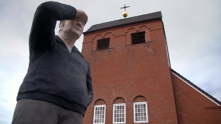 Lebensgroße Betonfigur eines Mannes, der sich den Arm über die Augen hält, im Hintergrund eine Kirche