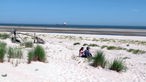 Zwei Personen sitzen am Sandstrand und schauen aufs Meer, wo ein großes Tankschiff entlangfährt