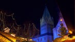 Blick über Dächer von Weihnachtsmarktbuden auf einen nächtlich beleuchteten Kirchturm