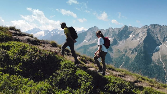 Peter Habeler und Tamina Kallert wandern auf einem Bergpfad vor Alpenkulisse