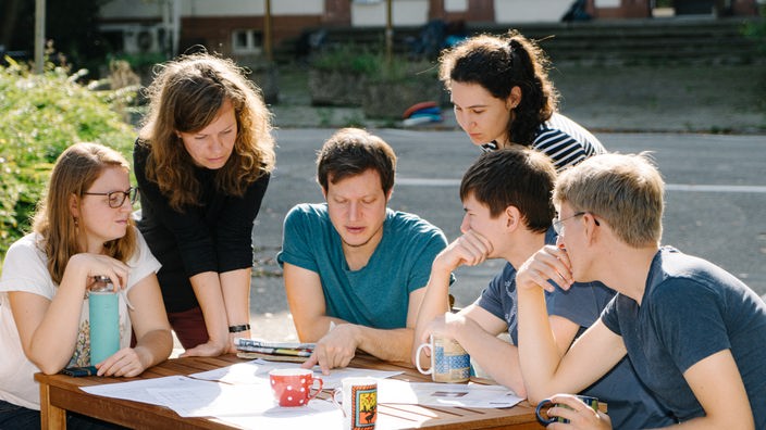 Eine Gruppe junger Menschen sitzt zusammen an einem Tisch und sind über nicht weiter erkennbare Pläne gebeugt. Sie trinken Kaffee.