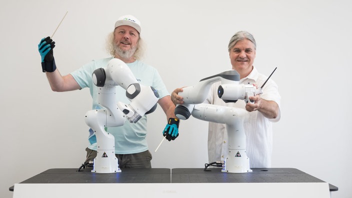Zwei Personen halten Taktstöck in den Händen, vor ihnen sind zwei Roboter-Arme aufgestellt.