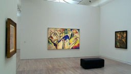 An einer weißen Museumswand hängt ein großes buntes und abstraktes Bild