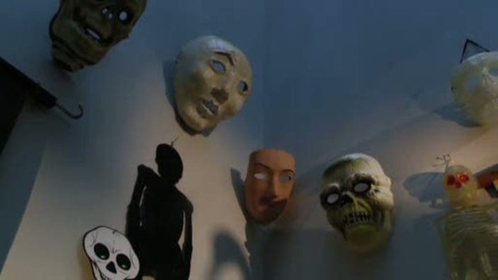 Verschiedene Masken, beispielsweise eine weiße, eine menschliche und eine Totenkopfmaske, hängen über Eck an einer Wand. Dazu verschiedene Skelettmodelle