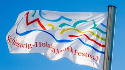 Eine Fahne mit dem Logo des Schleswig-Holstein Musik Festivals (SHMF) weht vor blauem Himmel.