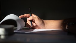 Eine Person hält einen Stift in der Hand und blättert ein Blatt Papier um.